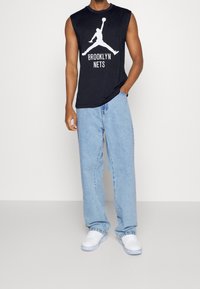 Man in een mouwloze zwarte Brooklyn Nets-shirt, losse lichtblauwe jeans en witte sneakers, staande met één hand in zijn zak.