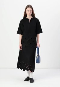 Svart klänning med korta ärmar, med utskurna blommönster i nederkanten. Modellen håller en blå handväska och bär svarta skor med vita strumpor.