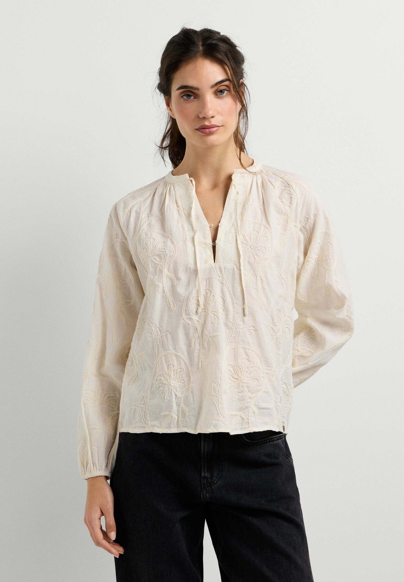 Jeune femme aux cheveux foncés portant un chemisier brodé crème à manches longues et un pantalon noir, debout devant un fond uni.