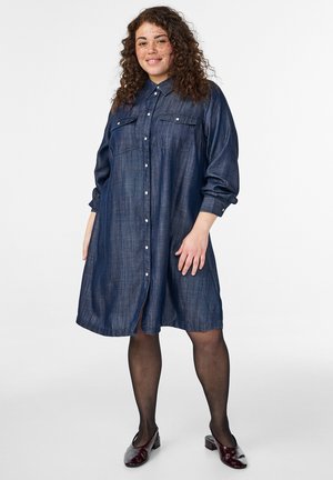 Naeratav naine lokkis juustega, kandes tumelilla triiksärgiga kleiti, musti sukkpükse ja burgundia madala kontsaga kingi, seisab lihtsa tausta ees.