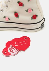 Kremfargede kanvas høy-top sneakers prydet med røde og rosa rose-applikasjoner. Inkluderer en rød hjerteformet tag med tekstene "Til" og "Fra".