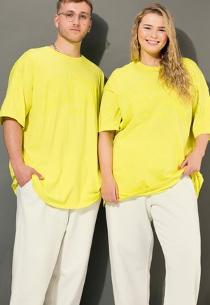 Dos adultos jóvenes con camisetas anchas de color amarillo brillante a juego y pantalones blancos, de pie frente a un fondo gris.