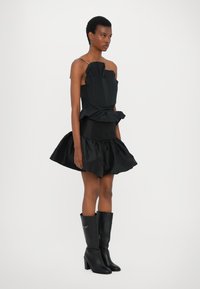 Black dress with layered ruffles, sleeveless design, and textured top. Paired with knee-high black heeled boots. Smooth fabric finish.