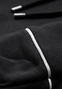 Zwarte hoodie stof die een close-up toont van een zilveren accent op de manchet, met een soepele textuur en subtiele stikselafwerking.