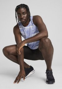 Sportowy top w jasnym niebieskim kolorze z poziomymi wzorami, zestawiony z czarnymi szortami i ciemnymi butami do biegania. Model w przysiadzie z skupionym wyrazem twarzy.