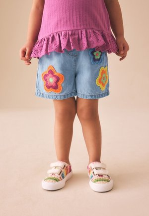 Barn iført lilla top, denimshorts med farverige blomsterlapper og multifarvede sneakers med velcrobånd på beige baggrund.