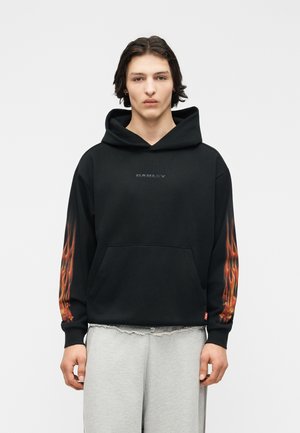 Giovane con capelli scuri che indossa una felpa nera con cappuccio decorata con fiamme sulle maniche e pantaloni grigio chiaro, in piedi davanti a uno sfondo semplice.