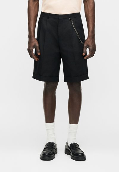 Άνδρας που φοράει μαύρα πιετέ σορτς με αλυσίδα, λευκές κάλτσες, μαύρα μοκασίνια και ανοιχτό μπεζ μπλουζάκι, στέκεται μπροστά σε απλό φόντο.