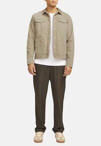 Veste en denim kaki avec col pointu, boutons avant, poches à rabat et t-shirt beige à col rond ; associé à un pantalon sombre et des baskets claires.