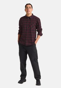 Chemise en flanelle à carreaux maroon et noir avec fermeture par boutons et poche poitrine, associée à un pantalon cargo noir et des chaussures noires.