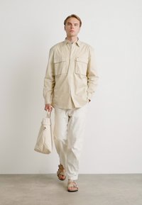 Beige, langetermijnshirt met lange mouwen en twee voorzakken, gecombineerd met lichte broeken en bruine sandalen. Houdt een bijpassende beige tote bag vast.