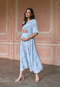 Gravid kvinde i en lyseblå blomsterkjole og hæle, der står indendørs og blidt holder om sin babybule og smiler blidt.