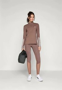 Brown athletic long-sleeve top with a half-zip and white stripes, matched with brown fitted leggings and white shoes, holding a black bag.