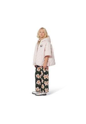 Jong meisje in een lichtroze gewatteerde hoodie en zwarte broek met grote roze en witte bloemmotieven, staand op een witte achtergrond.