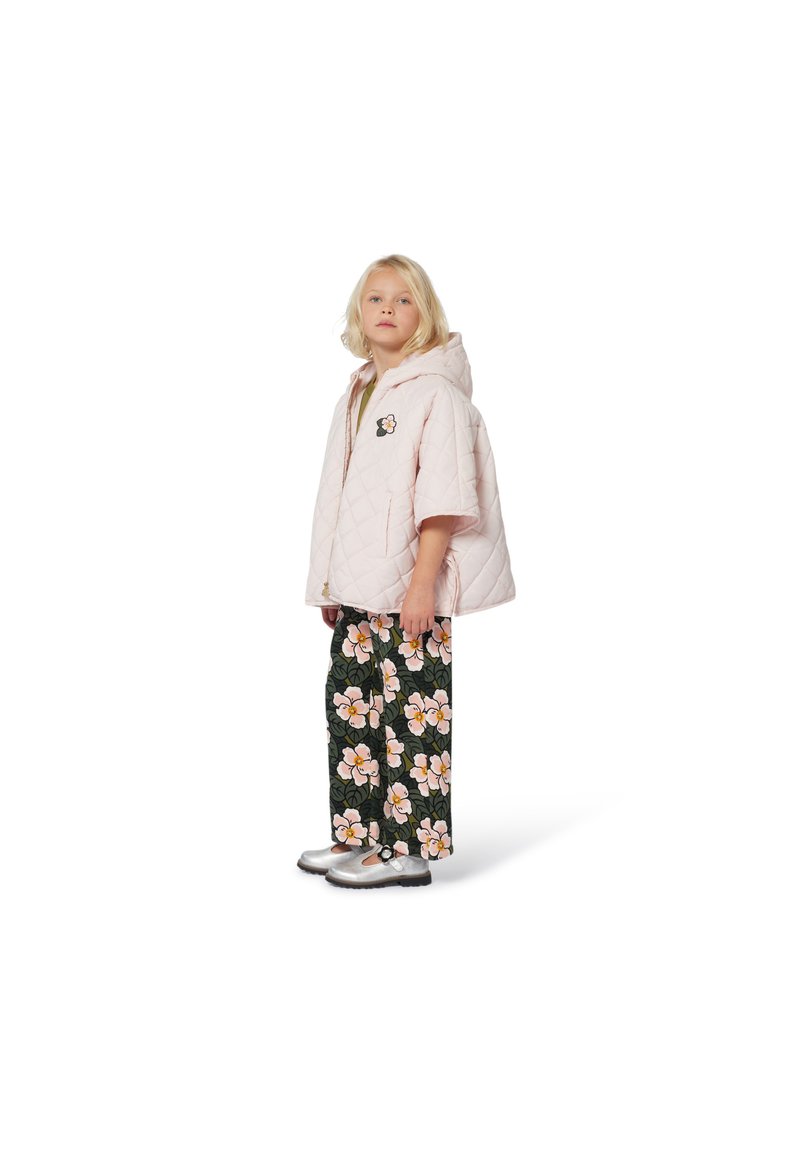 Jeune fille portant une veste à capuche matelassée rose pâle et un pantalon noir avec de grands motifs floraux roses et blancs, se tenant sur un fond blanc.