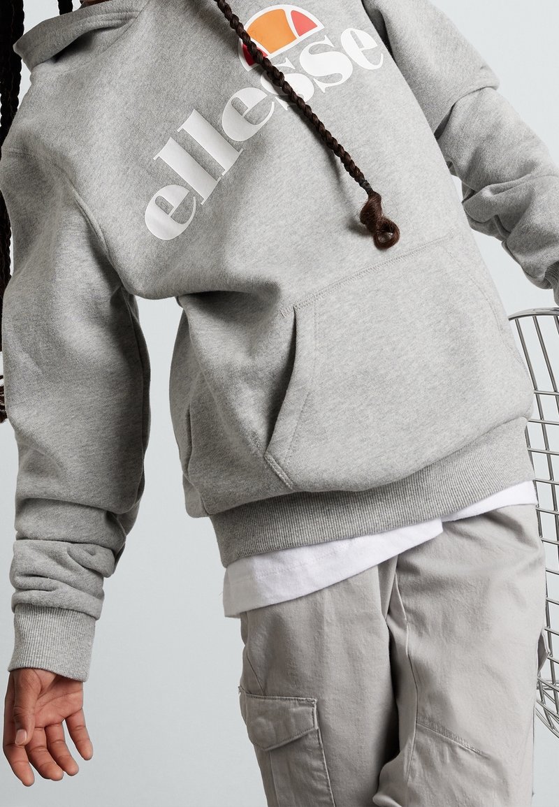 Sudadera gris con capucha y logo blanco de "ellesse" con acentos naranjas, que presenta un bolsillo tipo canguro y puños acanalados, usada sobre una camisa blanca.
