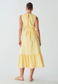 Keltainen hihaton mekko, jossa on istuva vyötärö, röyhelöhelma ja avara selkäosa, valmistettu kevyestä kankaasta. Sujuva pinta.