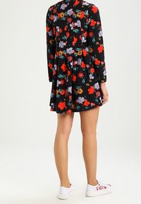 Schwarzes Blumenkleid mit langen Ärmeln, das mit roten, lila und orangen Blumen versehen ist. Kombiniert mit weißen Sneakers, die ein ausgestelltes Design zeigen.