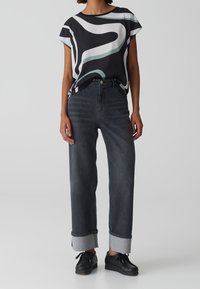 Kobieta ubrana w górę z abstrakcyjnym wzorem w czerni i bieli, ciemne, podwinięte dżinsy oraz czarne buty na sznurówki na jednolitym tle.