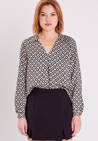 Blouse à manches longues en crème avec un motif en forme de losange noir, dotée d'un col en V et d'un léger tombé, associée à une jupe noire ajustée.
