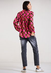 Langärmlige Bluse in Rosa und Schwarz mit geometrischem Diamantmuster, kombiniert mit ausgewaschenen grauen Jeans und weißen Sneakers. Rückansicht.
