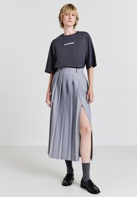 Jupe plissée grise avec une fente haute, associée à un t-shirt gris foncé arborant le texte blanc "PAS DE DRAME". Des mocassins noirs et des chaussettes grises viennent compléter le look.