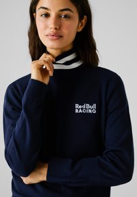 Pullover navy con un colletto alto a strisce e polsini a coste. Presenta il logo ricamato "Red Bull RACING" in bianco. Texture morbida.