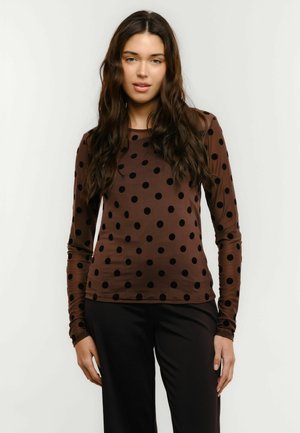 Tânără cu păr lung și închis la culoare, purtând o bluză maro cu mâneci lungi și buline negre, și pantaloni negri, stând în fața unui fundal simplu.