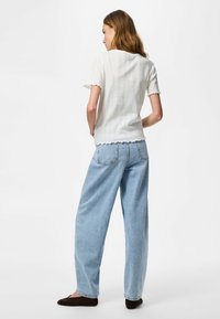 Hosszú hajú nő fehér rövid ujjú felsőben és világoskék, bő szárú farmernadrágban, keze a háta mögött, háttal áll.