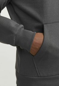 Charcoal szary sweter z prążkowanymi mankietami, kieszenią typu kangur i gładką teksturą. Ręka częściowo widoczna z ręką w kieszeni.