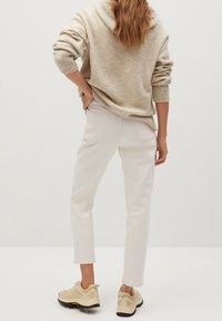 Persona vestida con un suéter beige, pantalones cortos off-white y zapatillas beige, de pie con una mano en la cadera, de espaldas al espectador.