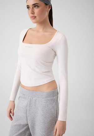 Femme portant un haut blanc ajusté à manches longues et un pantalon de survêtement gris, avec un bandeau gris, debout devant un fond clair uni.