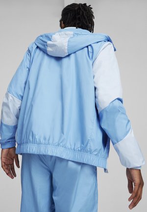 Chaqueta cortavientos de color azul claro con capucha, que presenta acentos en blanco contrastantes en las mangas y una cintura elástica para un ajuste seguro.