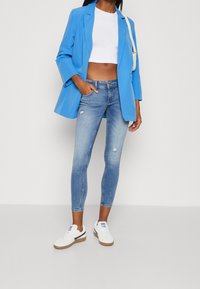 Blazer azul sobre un top corto blanco, combinado con jeans ajustados rasgados de color azul claro y zapatillas blancas con acentos oscuros.