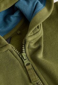 Veste à capuche zippée olive avec une doublure intérieure bleue. Elle dispose d'une fermeture éclair à l'avant et d'un tissu doux et texturé. Les accents comprennent des coutures visibles.