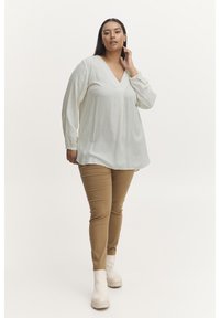 Blouse en crème clair avec un col en V, des manches longues et des points texturés, associée à un pantalon beige ajusté et des bottines blanches cassées.