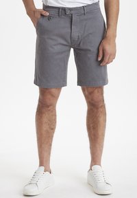 Pantalones cortos de algodón gris con un frente plano, dos bolsillos laterales y cierre de botón. Llevados con zapatillas blancas sobre un fondo liso.