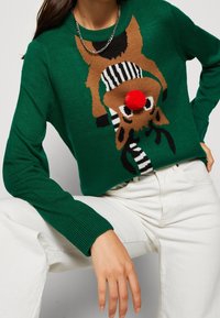 Grüner Strickpullover mit einem Rentierdesign, das eine rote Pompon-Nase und schwarz-weiße Streifen aufweist, kombiniert mit weißen Hosen.
