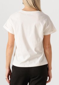 Vrouw die een effen wit kortärmlig T-shirt en zwarte broek draagt, gefotografeerd van achteren tegen een neutrale achtergrond.