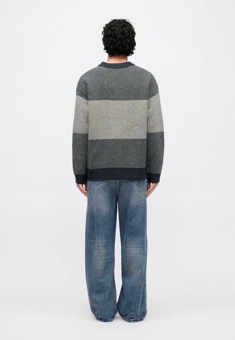 Pull en maille à rayures bleu marine, crème et gris avec un col rond ; associé à un jean bleu délavé à coupe ample. Tissu texturé sur toute la surface.