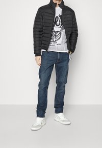 Černá péřová bunda s horizontálním prošíváním, šedá mikina s grafikou, modré džíny a bílé tenisky.