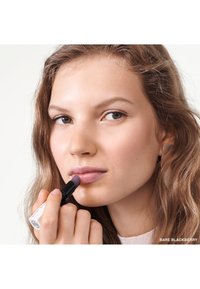 Rouge à lèvres dans un tube noir avec une marque blanche, appliqué sur les lèvres. Le modèle a des cheveux ondulés, une peau douce et porte une tenue rose clair.