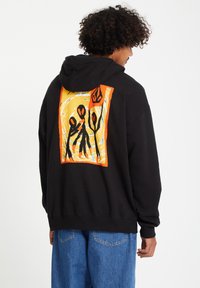Zwarte hoodie met een kleurrijke achterafbeelding van abstracte figuren en bloemen op een gele achtergrond met oranje accenten.