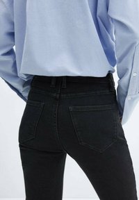 Los jeans negros de cintura alta presentan un corte ajustado, diseño de cinco bolsillos y una tela de mezclilla texturizada. Una camisa de color azul claro está metida en la cintura.