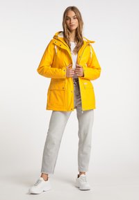 Giacca impermeabile gialla con cappuccio, lacci e accessori in argento. Presenta due tasche frontali e un interno foderato in pile. Abbinata a jeans grigi e scarpe da ginnastica bianche.
