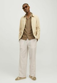 Chaqueta de lino beige claro, camisa marrón de botones, pantalones de textura clara y zapatos marrones de tipo slip-on. Los accesorios incluyen gafas de sol.