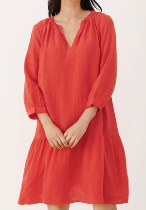 Vrouw in een loszittende, lange mouwen, knielange rode jurk met geplooide halslijn en manchetten, staand met samengevouwen handen voor zich.