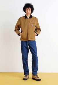Chaqueta marrón con cremallera y cuello negro, vaqueros azules y botas de tonos marrones. La chaqueta tiene un bolsillo frontal con un parche de logo.