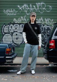 Μαύρο T-shirt πάνω από ένα λευκό μακρυμάνικο πουκάμισο, ανοιχτό μπλε φαρδιά τζιν, λευκά αθλητικά παπούτσια με μαύρες λεπτομέρειες, γκρι καπέλο μπέιζμπολ, τσαντάκι ώμου.