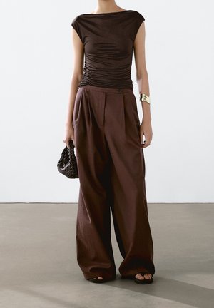 Haut brun ajusté à manches courtes avec une texture froncée associé à un pantalon large et fluide brun. Accessoirisé avec un sac tissé noir et un bracelet en or.